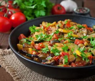 salteado de verduras sobre un plato negro hondo en un ambiente rústico con tomates y perejil al fondo