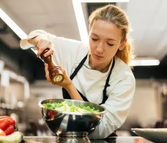 mujer chef salpimentando una ensalada sobre un recipiente metálico en una cocina profesional