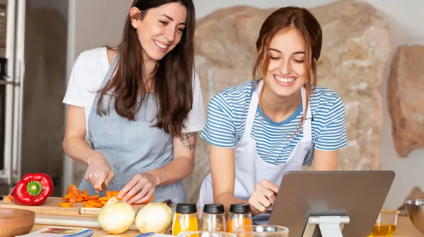 dos chicas sonrientes frente a unos ingredientes mirando una pantalla y cortando zanahorias